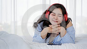 Asian young happy calm fat overweight woman lay down close eyes smiling peacefully on white sheet blanket listening to music from