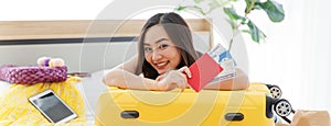 Asian young happy female traveler showing red passport with air tickets leaning on big yellow trolley luggage on bed look at