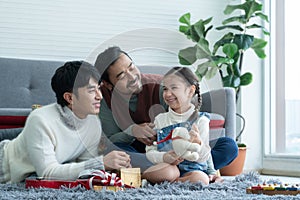 Asian young LGBTQ gay couple giving gift to little Caucasian adopted kid in living room at home. Kid smiling and delighted