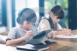 Asian young little children self learn on table in living room at home. Cute Boy use tablet and headphone for E-learning and girl