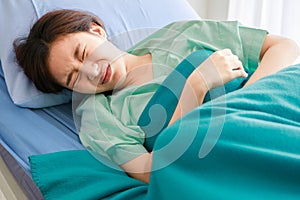 Asian young long hair sick female patient wears green hospital uniform lay down on bed covered by blanket hold hands on stomach