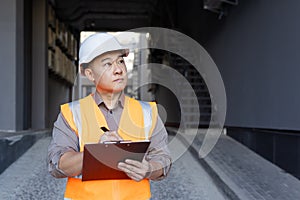 Asian young man builder, expert and inspector inspects and records work progress. Standing in a hard hat and vest