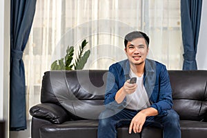 Asian young man holding remote control and watching TV while sitting on the couch at home