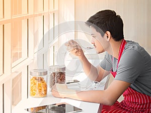 Asian young man sitting next to window glass and having cereal with milk on table for breakfast at home in the morning.