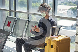 Asian young man traveller new normal wearing face mask sitting Social distancing holding smartphone in Airplane lounge. New normal