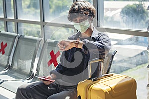 Asian young man traveller new normal wearing face mask sitting Social distancing holding smartphone look at wristwatch in Airplane