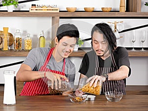 Asian young men wear apron are pouring cereal into a glass bowl together on wooden table.