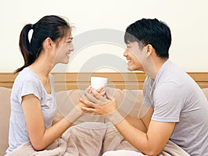 Asian young man and woman holding coffee cup on the bed. The lover smile and look at each other face happily