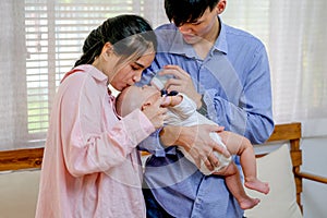 Asian young mother kiss her little baby drink milk from bottle and is held by his father and they stand in living room with soft