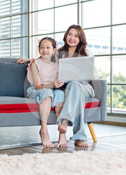 Asian young mother sitting on cozy sofa couch in living room pointing teaching little girl daughter using notebook computer