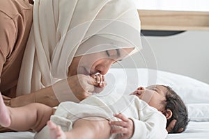 Asian young Muslim mother wearing hijab kissing hand of her newborn baby 1 month old while cute infant grabbing mom finger,