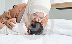 Asian young Muslim mother wearing hijab kissing her newborn baby 1 month old on forehead while cute infant sleeping on bed in