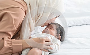 Asian young Muslim mother wearing hijab kissing her smiling newborn baby 1 month old on forehead while cute infant sleeping on bed