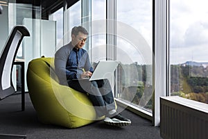 Asian young office worker sitting on soft ottoman chair, programmer using laptop for writing code and programming