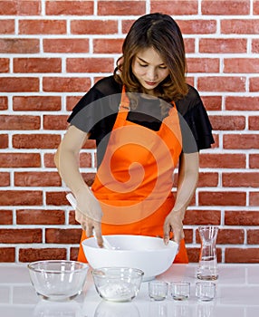 Asian young pastry baker in orange apron stand pouring water from glass in white ceramic bowl of flour mixing dough with plastic