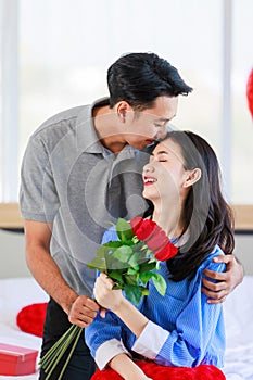 Asian young romantic cheerful lover couple male boyfriend sitting on bed giving red roses bouquet kissing surprise female