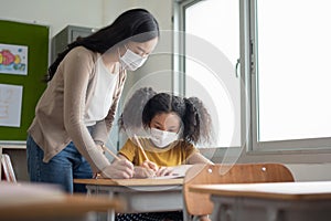 Asian Young Teacher teaching her African American student girl in protective face mask in classroom