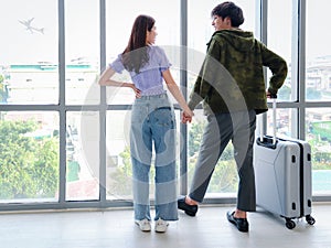 Asian young tourist lover couple hold each other hand, looking through window, standing with luggage suitcase from behind, waiting