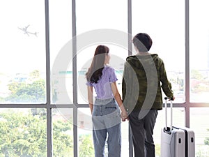 Asian young tourist lover couple hold each other hand, looking through window, standing with luggage suitcase from behind, waiting