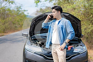 Asian young unhappy man talking on a cell phone  in front of the open hood  broken down car On Country Road Phoning For Help.