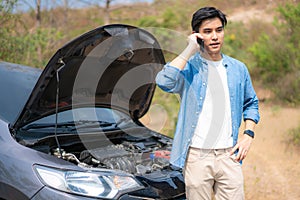 Asian young unhappy man talking on a cell phone  in front of the open hood  broken down car On Country Road Phoning For Help.