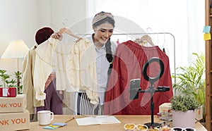 Asian young woman blogger showing clothes in front of smartphone camera while recording vlog video and live streaming at her shop