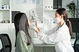 Asian young woman doctor using flashlight on patient`s eye in hospital. Attractive specialist physician wear mask, examining and