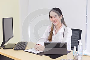 Asian young woman doctor in a white lab coat is sitting on office room at hospital while has a computer, clipbroad and alcohol gel