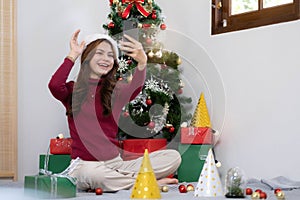 asian young woman has video chat on smartphone and shows present box to her friend with christmas tree