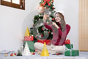 asian young woman has video chat on smartphone and shows present box to her friend with christmas tree