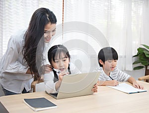 Asian young woman help her son do homework with daughter using laptop sit beside on table in living room at home