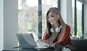asian young woman listening music with headphone and streaming music from laptop on sofa relaxing at home