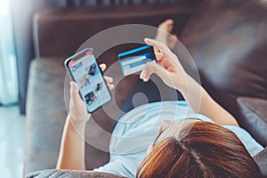 Asian young woman lying down on sofa resting browsing internet with Smart Phone holding credit card online shopping concept