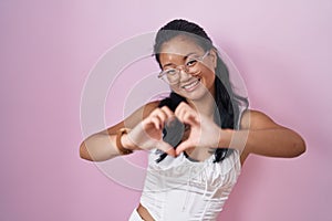 Asian young woman standing over pink background smiling in love showing heart symbol and shape with hands