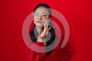 Asian young woman standing over red background hand on mouth telling secret rumor, whispering malicious talk conversation
