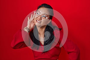 Asian young woman standing over red background smiling with hand over ear listening an hearing to rumor or gossip