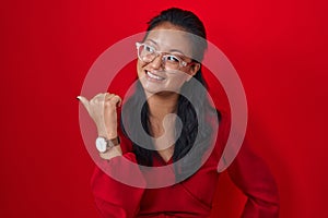 Asian young woman standing over red background smiling with happy face looking and pointing to the side with thumb up