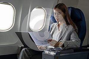 Asian young woman using laptop with financial report sitting near windows at first class on airplane during flight