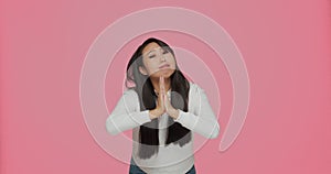 Asian young woman wishing something with folded hands asking help or forgiveness, pleading on pink studio background