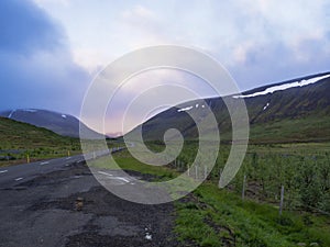 Asphalt road curve through empty northern landscape with green grass colorful hills and sunset dramatic sky, way to the