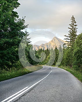 Asphalt road in forest, coniferous trees on both sides, mount Krivan peak Slovak symbol with afternoon sun clouds, in