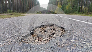 The asphalt road with markings has a damaged surface and a pothole with small stones and pieces of asphalt in it. The road passes