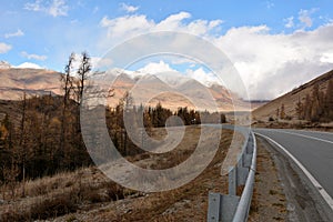 An asphalt road with metal fences descends from the mountain into a picturesque autumn valley at the foot of snow-capped mountains