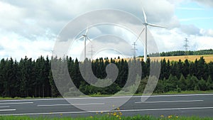 Asphalt road with riding motor bikers and Blades of wind turbines rotating under cloudy sky in back