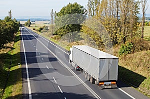 Asphalt road between the trees in the countryside. Two white trucks on the road.