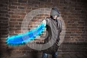 Assasin stands in a cloak with a hood covering his eyes and holds a glowing blue sword in front of him with one hand. Selective