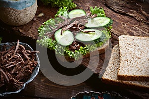 Assembling sandwich with meat, dark bread, fresh green salad, dry tomatoes and fresh cucumbers on rustic wooden table surface