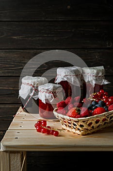 Assorted berries in a basket, glass jars with jams from different berries on wooden table