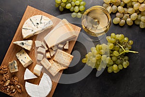 Assorted cheeses with white grapes, walnuts, crackers and white wine on a wooden Board. Food for a romantic date on a dark stone b