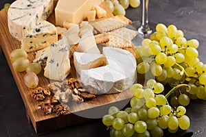 Assorted cheeses with white grapes, walnuts, crackers and white wine on a wooden Board. Food for a romantic date on a dark stone b